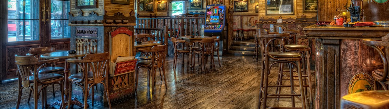 Na zdjęciu jest wnętrze portowego baru ze stolikami z krzesłami, hokery, bar, na ścianach wiszą obrazy o tematyce morskiej